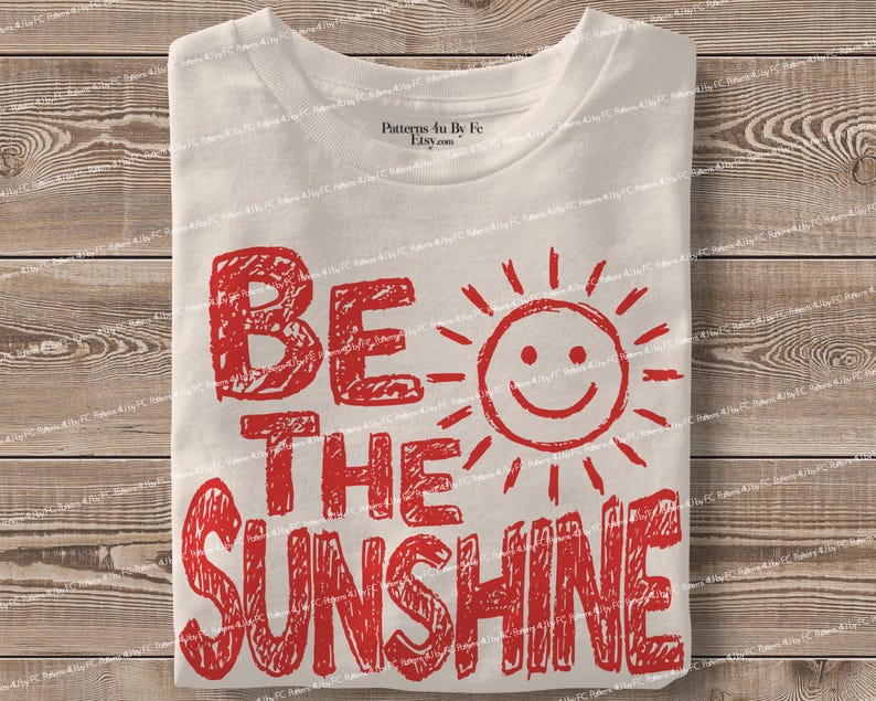May include: A folded white t-shirt with the red text "BE THE SUNSHINE" and a smiling sun graphic. The shirt is made of a soft material and is folded neatly on a wooden surface.