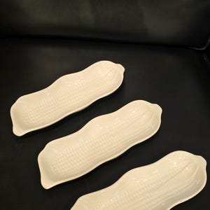 May include: Three off-white ceramic corn-shaped serving dishes. Each dish has a textured surface mimicking corn kernels and a gently curved shape. The dishes are arranged on a dark surface.