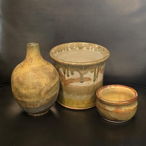 May include: Three handcrafted ceramic pottery pieces in earthy tones. A bulbous vase, a cylindrical pot with dripped glaze, and a small bowl. The pottery features shades of brown, tan, and green, with a rustic, textured appearance.