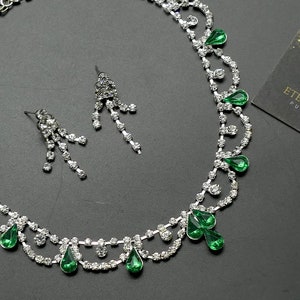 May include: A silver necklace and earring set with clear and emerald green rhinestones. The necklace has a delicate design with teardrop-shaped green stones. The earrings are dangling with multiple clear rhinestones. A card with the text "Eternal Sparkle Pure Elegance" is in the background.