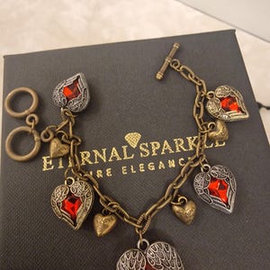 May include: A vintage bracelet with heart-shaped charms and a toggle clasp. The bracelet features red gemstone hearts, small gold-tone hearts, and a larger heart pendant with wing details. The bracelet is on a dark grey box with the text "Eternal Sparkle".