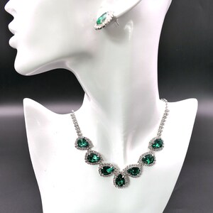 Puede incluir: Un conjunto de collar y pendientes plateados sobre una cabeza de maniqu&iacute; blanca. La joyer&iacute;a presenta piedras verdes esmeralda en forma de l&aacute;grima rodeadas de cristales transparentes. Los pendientes son un par a juego, y el collar tiene una cadena delicada.