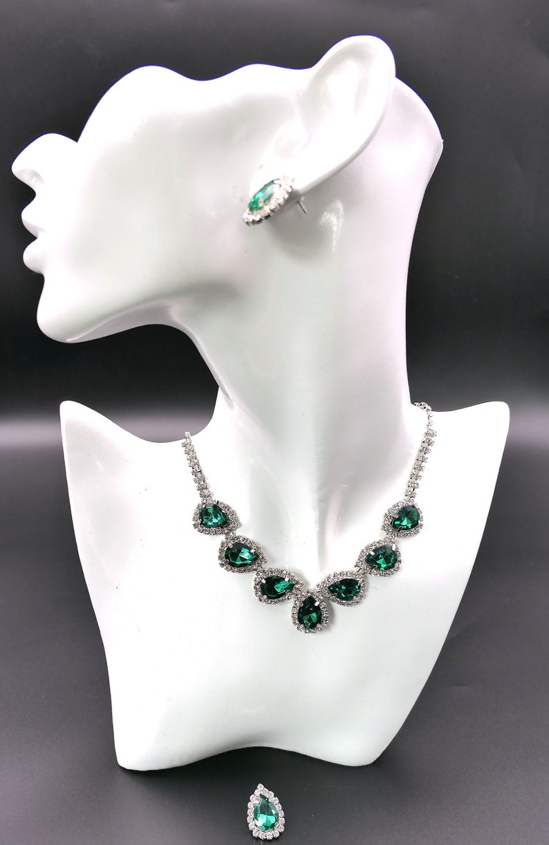 Puede incluir: Un conjunto de collar y pendientes plateados sobre una cabeza de maniqu&iacute; blanca. La joyer&iacute;a presenta piedras verdes esmeralda en forma de l&aacute;grima rodeadas de cristales transparentes. Los pendientes son un par a juego, y el collar tiene una cadena delicada.