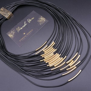 May include: A multi-strand black necklace with gold-colored curved tube accents. The necklace has a magnetic clasp and is displayed with a small card that reads "Thank You" and "Eternal Sparkle, Pure Elegance".