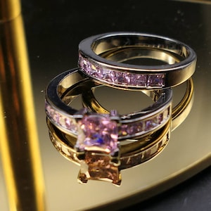 Peut inclure: Deux bagues, une alliance et une solitaire, reposent sur une surface réfléchissante. Les bagues ont un anneau argenté avec un intérieur doré. L'alliance est ornée de petites pierres roses, tandis que la solitaire présente une grosse pierre rose taillée en carré.