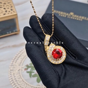 May include: Gold necklace with a pendant. The pendant has a large, round red gemstone encircled by smaller clear stones. The chain is delicate. The text "Eternal Sparkle" is visible.