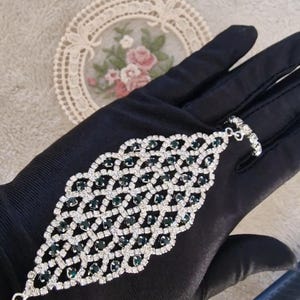 Puede incluir: Una pulsera plateada con un diseño geométrico, con brillantes transparentes y verde esmeralda. La pulsera se muestra sobre una superficie negra, resaltando su intrincado patrón y detalles brillantes. Esta pieza de joyería es elegante y llamativa.