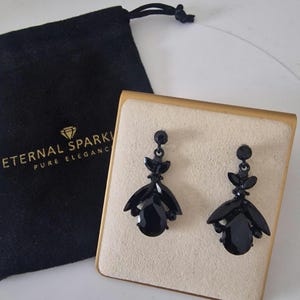 May include: A pair of black drop earrings featuring faceted teardrop and marquise-shaped stones, displayed on a beige square card. Beside the earrings is a black drawstring pouch with the gold text ETERNAL SPARKLE PURE ELEGANCE printed on it.