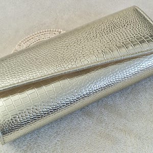 May include: Silver clutch bag with a textured, crocodile-effect pattern. The rectangular bag has a fold-over flap and a shiny finish. The clutch is likely made of a synthetic material and is suitable for formal events.