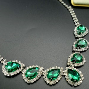 Puede incluir: Un collar brillante con grandes piedras preciosas verdes esmeralda en forma de l&aacute;grima. Cada gema est&aacute; rodeada por un halo de peque&ntilde;os cristales transparentes, engastados en metal plateado. Dise&ntilde;o cl&aacute;sico y elegante.