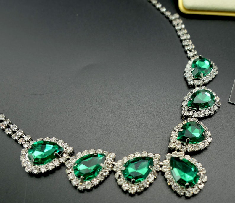 Puede incluir: Un collar brillante con grandes piedras preciosas verdes esmeralda en forma de l&aacute;grima. Cada gema est&aacute; rodeada por un halo de peque&ntilde;os cristales transparentes, engastados en metal plateado. Dise&ntilde;o cl&aacute;sico y elegante.