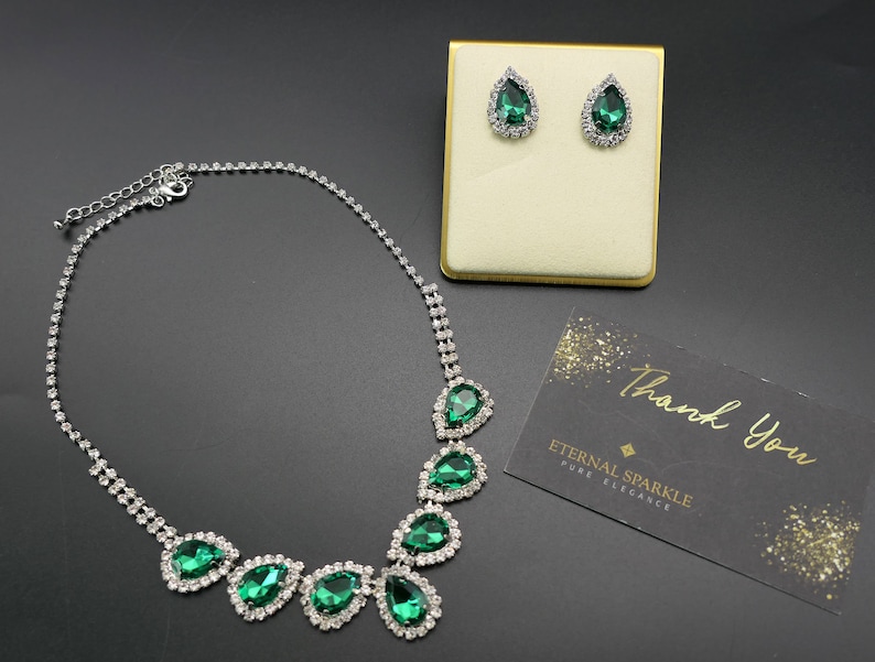 Puede incluir: Un conjunto de collar y pendientes plateados con piedras en forma de l&aacute;grima verde esmeralda y brillantes transparentes. El conjunto se exhibe sobre una superficie negra con un expositor dorado y una tarjeta de agradecimiento.