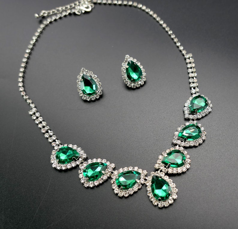 Puede incluir: Un conjunto de collar y pendientes plateados. El collar presenta piedras en forma de l&aacute;grima de color verde esmeralda, cada una rodeada de cristales transparentes m&aacute;s peque&ntilde;os. Los pendientes repiten el dise&ntilde;o con una sola piedra en forma de l&aacute;grima.