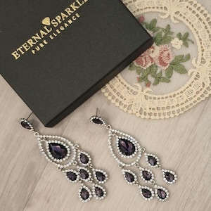 Peut inclure: Boucles d'oreilles pendantes argentées ornées de pierres en forme de goutte de couleur violet foncé et de cristaux transparents. Elles sont présentées à côté d'une boîte noire avec le texte doré "ETERNAL SPARKLE PURE ELEGANCE".