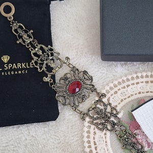 Può includere: Un braccialetto color oro antico con un design ornato e una gemma rossa ovale centrale. Il braccialetto è esposto con un sacchetto di velluto nero e una scatola grigio scuro. I dettagli intricati e l'estetica vintage del braccialetto sono evidenziati.