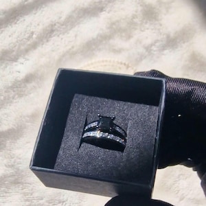 May include: A black ring set in a black box. The engagement ring features a square, black centre stone, and the band is encrusted with small, clear stones. The matching band also has small, clear stones. The box is held by a person wearing a black glove.