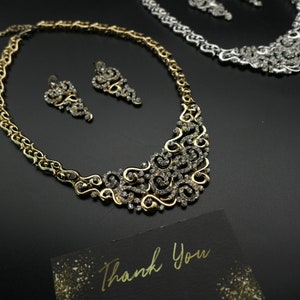 May include: A gold necklace and earrings set with a delicate, ornate design featuring clear rhinestones. The necklace has a crescent shape and the earrings are dangling.