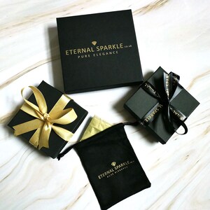 Puede incluir: Vista cenital de varias cajas de regalo negras y una bolsa negra con cord&oacute;n, todas con texto y detalles dorados. Las cajas est&aacute;n atadas con cintas doradas y negras. El texto dice "ETERNAL SPARKLE.co.uk PURE ELEGANCE."
