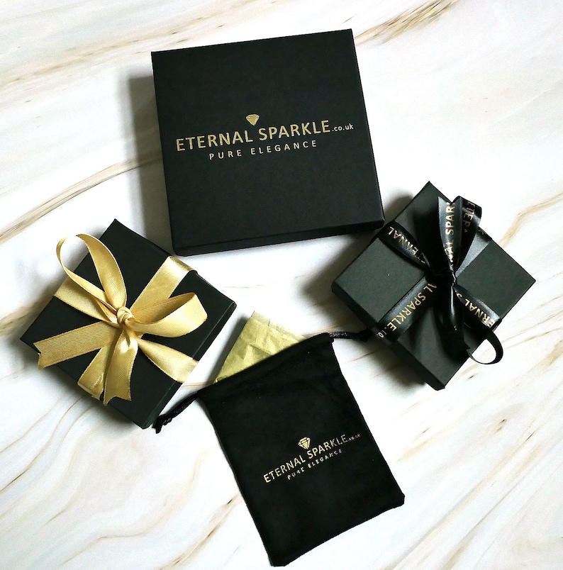 Puede incluir: Vista cenital de varias cajas de regalo negras y una bolsa negra con cord&oacute;n, todas con texto y detalles dorados. Las cajas est&aacute;n atadas con cintas doradas y negras. El texto dice "ETERNAL SPARKLE.co.uk PURE ELEGANCE."