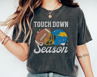Puede incluir: Camiseta gris oscuro con la frase "Touch Down Season" sobre un balón de fútbol americano y un casco azul. El balón es marrón y el casco es azul y amarillo. La camiseta se lleva con vaqueros azules y zapatillas blancas.
