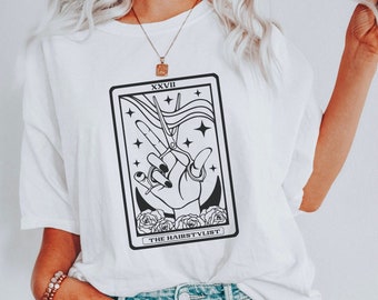 May include: White t-shirt with a black and white tarot card design featuring a hand holding scissors and the text "XXVIII THE HAIRSTYLIST".