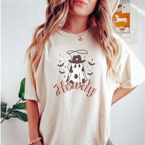 May include: A cream colored t-shirt with a cartoon ghost wearing a cowboy hat and a lasso. The ghost has black and white cow spots. The text "Howdy" is written in pink below the ghost.