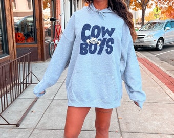 May include: A gray hooded sweatshirt with the text "COWBOYS" in blue lettering with a cowboy hat graphic. The model is wearing the sweatshirt with white sneakers.