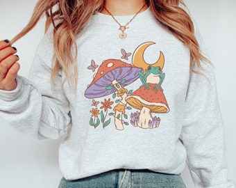 May include: A gray sweatshirt with a graphic of a frog sitting on a mushroom with a crescent moon in the background. The mushroom is surrounded by flowers and crystals.