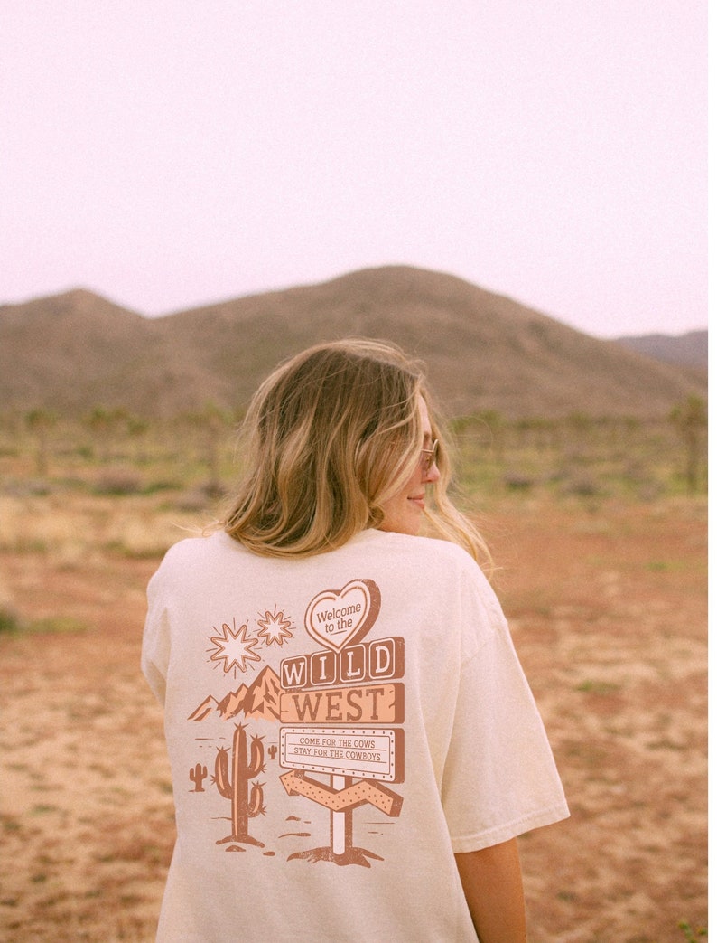 May include: A white t-shirt featuring a brown graphic print of a sign that reads "Welcome to the Wild West" with a cactus and mountains in the background. The sign also says "Come for the cows, stay for the cowboys".