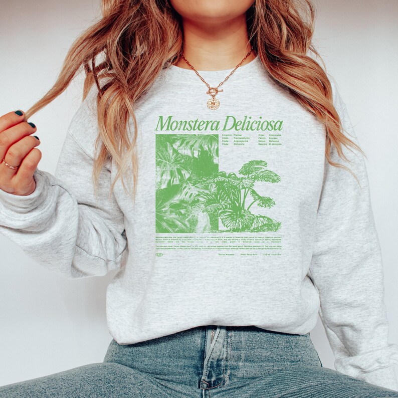 May include: A gray sweatshirt with a green graphic print of a Monstera Deliciosa plant. The graphic includes the plant's scientific name, origin, family, order, and other details. The text "Monstera Deliciosa" is printed in large, bold letters at the top of the graphic.