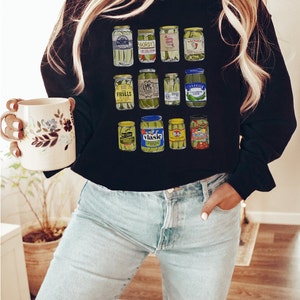 Op de afbeelding: Een zwart sweatshirt met een grafische print van verschillende augurkenpotten. De potten zijn in een rasterpatroon gerangschikt en tonen verschillende augurkenmerken, waaronder Gordy's, Vlasic en B&G.