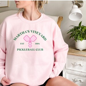 May include: Pink sweatshirt with a green graphic that says "Martha's Vineyard Est 1994 Pickleball Club" with a pink pickleball paddle and ball.
