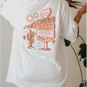 May include: White oversized t-shirt featuring a graphic print of a sign that reads "Welcome to the Wild West" with a heart, cactus, and mountains. The sign also says "Come for the cows, stay for the cowboys."
