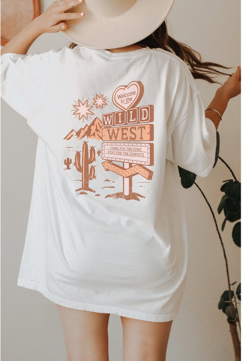 May include: White oversized t-shirt featuring a graphic print of a sign that reads "Welcome to the Wild West" with a heart, cactus, and mountains. The sign also says "Come for the cows, stay for the cowboys."