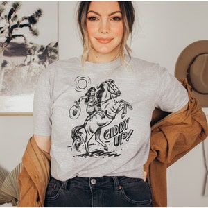 Peut inclure: Un t-shirt gris avec une illustration en noir et blanc d'une femme &agrave; cheval. Le texte "Giddy Up!" est imprim&eacute; sous l'illustration.