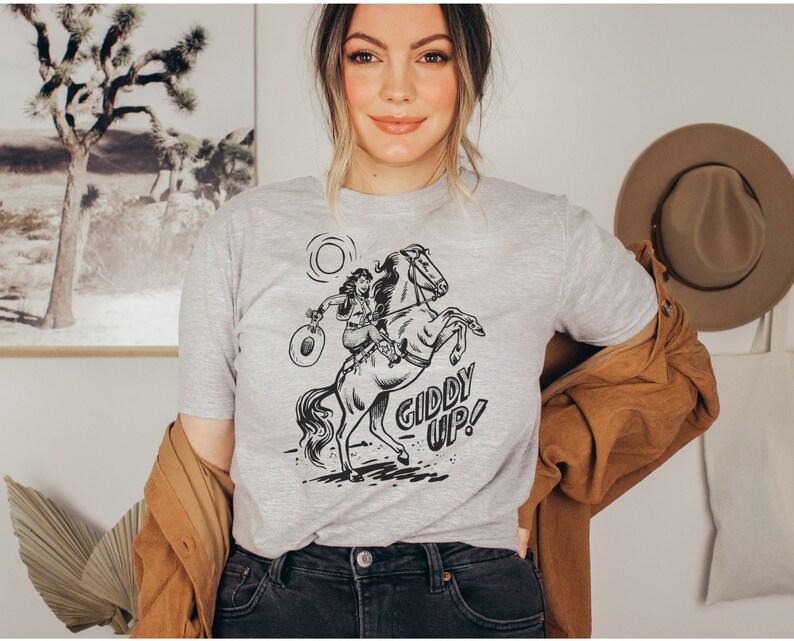 Peut inclure: Un t-shirt gris avec une illustration en noir et blanc d'une femme &agrave; cheval. Le texte "Giddy Up!" est imprim&eacute; sous l'illustration.