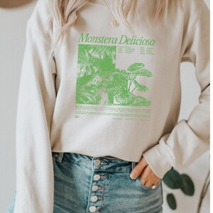 May include: A light beige sweatshirt with a green graphic print of a Monstera Deliciosa plant and text that reads "Monstera Deliciosa". The sweatshirt has a crew neck and long sleeves.