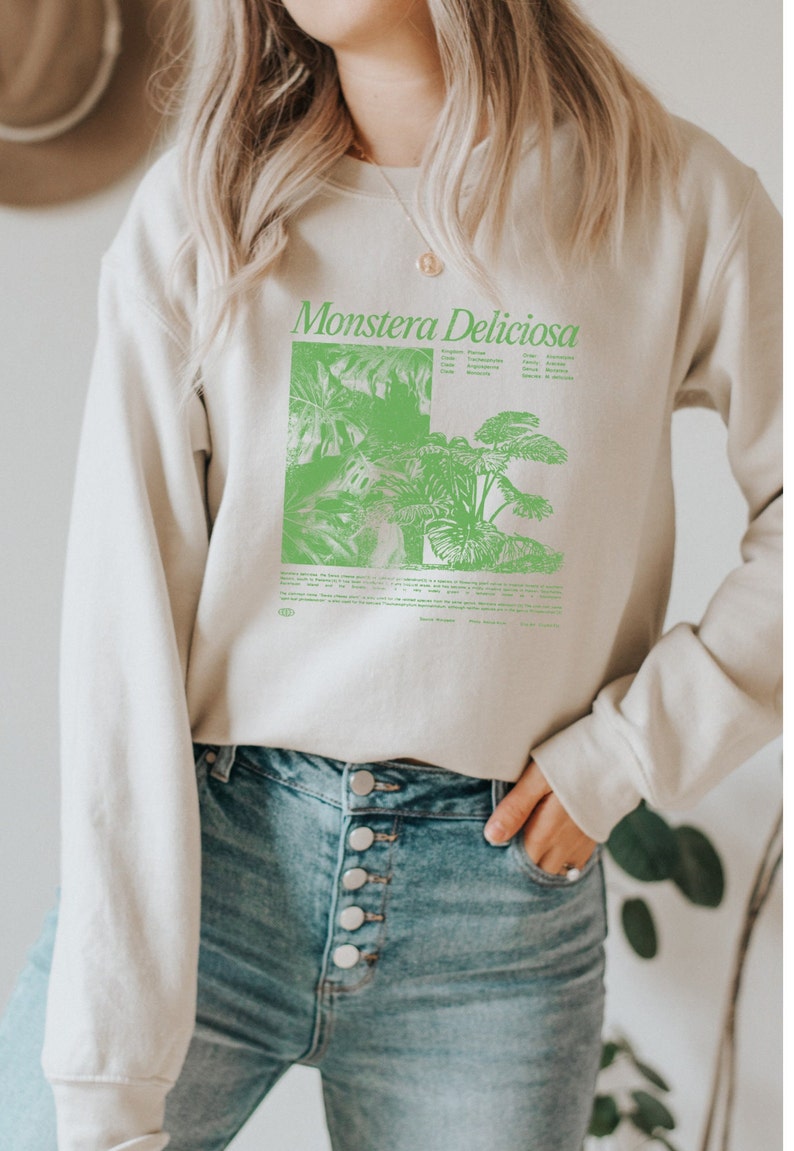 May include: A light beige sweatshirt with a green graphic print of a Monstera Deliciosa plant and text that reads "Monstera Deliciosa". The sweatshirt has a crew neck and long sleeves.