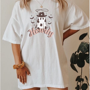 May include: White oversized t-shirt with a graphic of a ghost wearing a cowboy hat and the word "Howdy" printed in pink. The ghost has black and white cow spots.