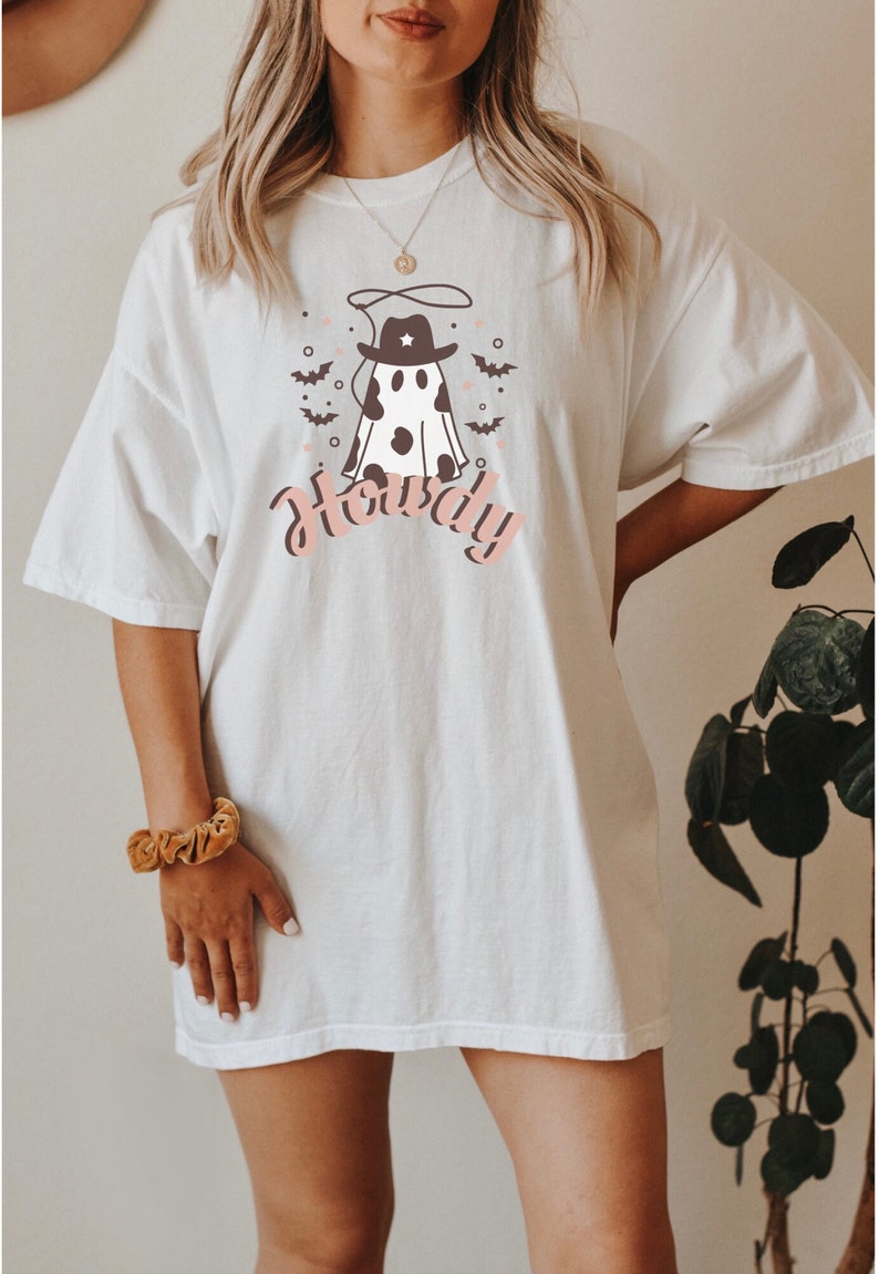May include: White oversized t-shirt with a graphic of a ghost wearing a cowboy hat and the word "Howdy" printed in pink. The ghost has black and white cow spots.