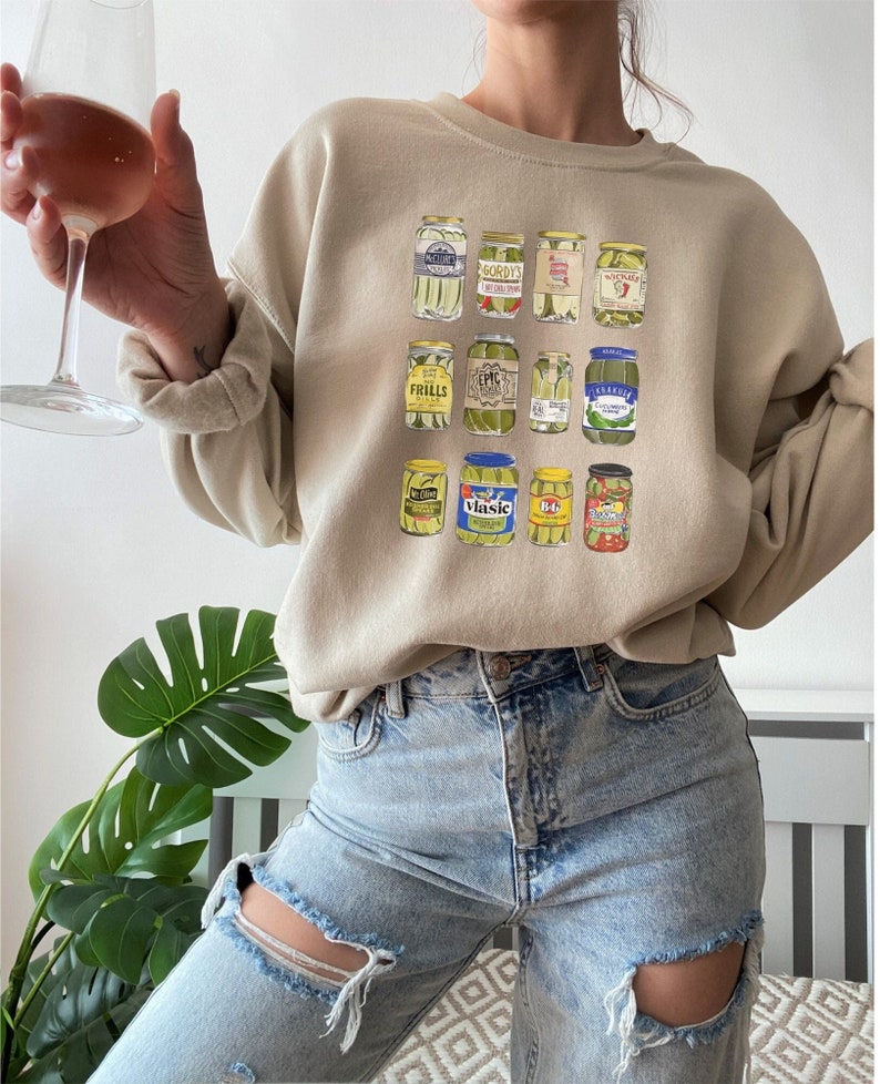 Op de afbeelding: Een beige sweatshirt met een grafische print van verschillende potten augurken. De potten hebben verschillende merknamen en labels, waaronder "Gordy's", "Frills", "Epic", "Vlasic" en "B.G.".