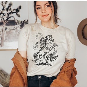 Peut inclure: Un t-shirt blanc avec une illustration en noir et blanc d'une femme &agrave; cheval. Le texte "Giddy Up!" est imprim&eacute; sous l'illustration.