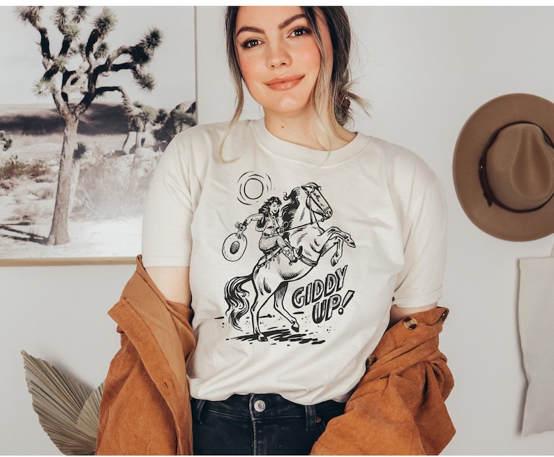 Peut inclure: Un t-shirt blanc avec une illustration en noir et blanc d'une femme &agrave; cheval. Le texte "Giddy Up!" est imprim&eacute; sous l'illustration.