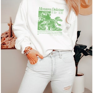 May include: White sweatshirt with a green illustration of a Monstera Deliciosa plant and the text "Monstera Deliciosa" printed on the front. The sweatshirt is being worn by a person with long blonde hair and is paired with light blue jeans.