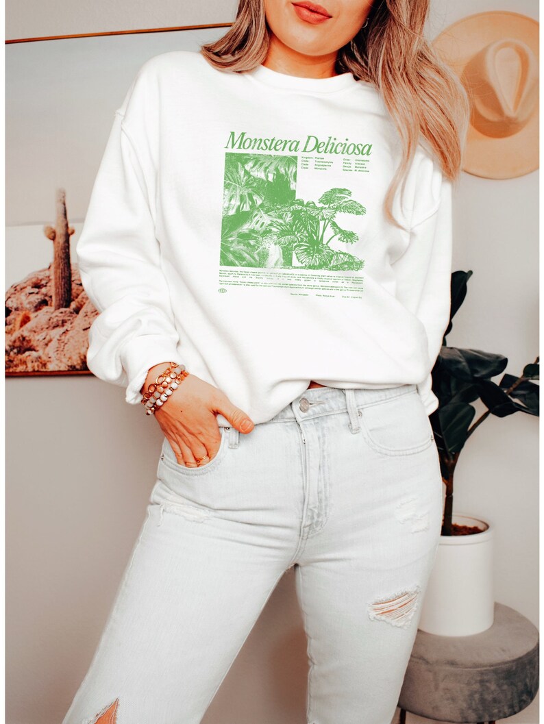 May include: White sweatshirt with a green illustration of a Monstera Deliciosa plant and the text "Monstera Deliciosa" printed on the front. The sweatshirt is being worn by a person with long blonde hair and is paired with light blue jeans.