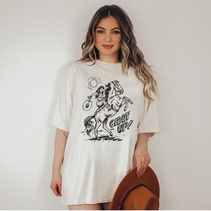Peut inclure: Un t-shirt blanc surdimensionn&eacute; avec un motif noir et blanc d'une femme &agrave; cheval. Le motif comprend le texte "Giddy Up!"