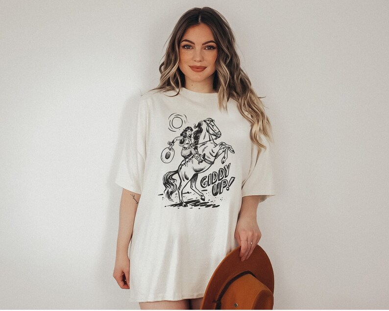 Peut inclure: Un t-shirt blanc surdimensionn&eacute; avec un motif noir et blanc d'une femme &agrave; cheval. Le motif comprend le texte "Giddy Up!"