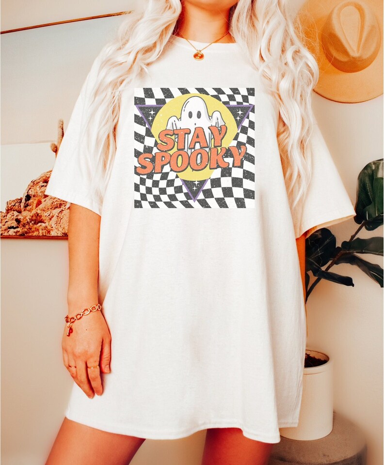 May include: White oversized t-shirt with a graphic of a ghost in a yellow circle with the text "STAY SPOOKY" in orange and purple letters on a black and white checkered background.