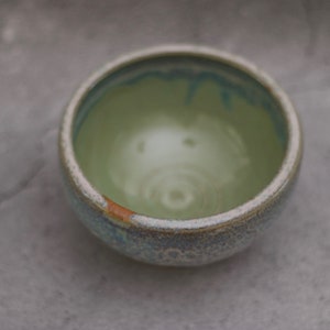 May include: A small, light green ceramic bowl with a light blue and brown glaze. The bowl has a slightly textured surface and is filled with a light green liquid.