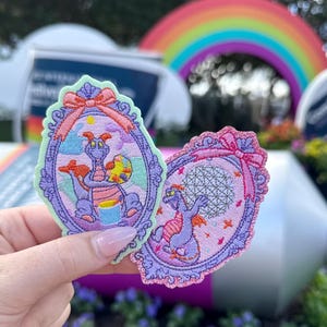 Könnte beinhalten: Zwei gestickte Aufnäher mit einer lila Drachenfigur. Einer zeigt den Drachen mit einer Farbpalette, der andere mit einem Globus. Beide sind in verzierten, ovalen Rahmen mit Schleifen eingefasst. Im Hintergrund ist ein Regenbogen zu sehen.